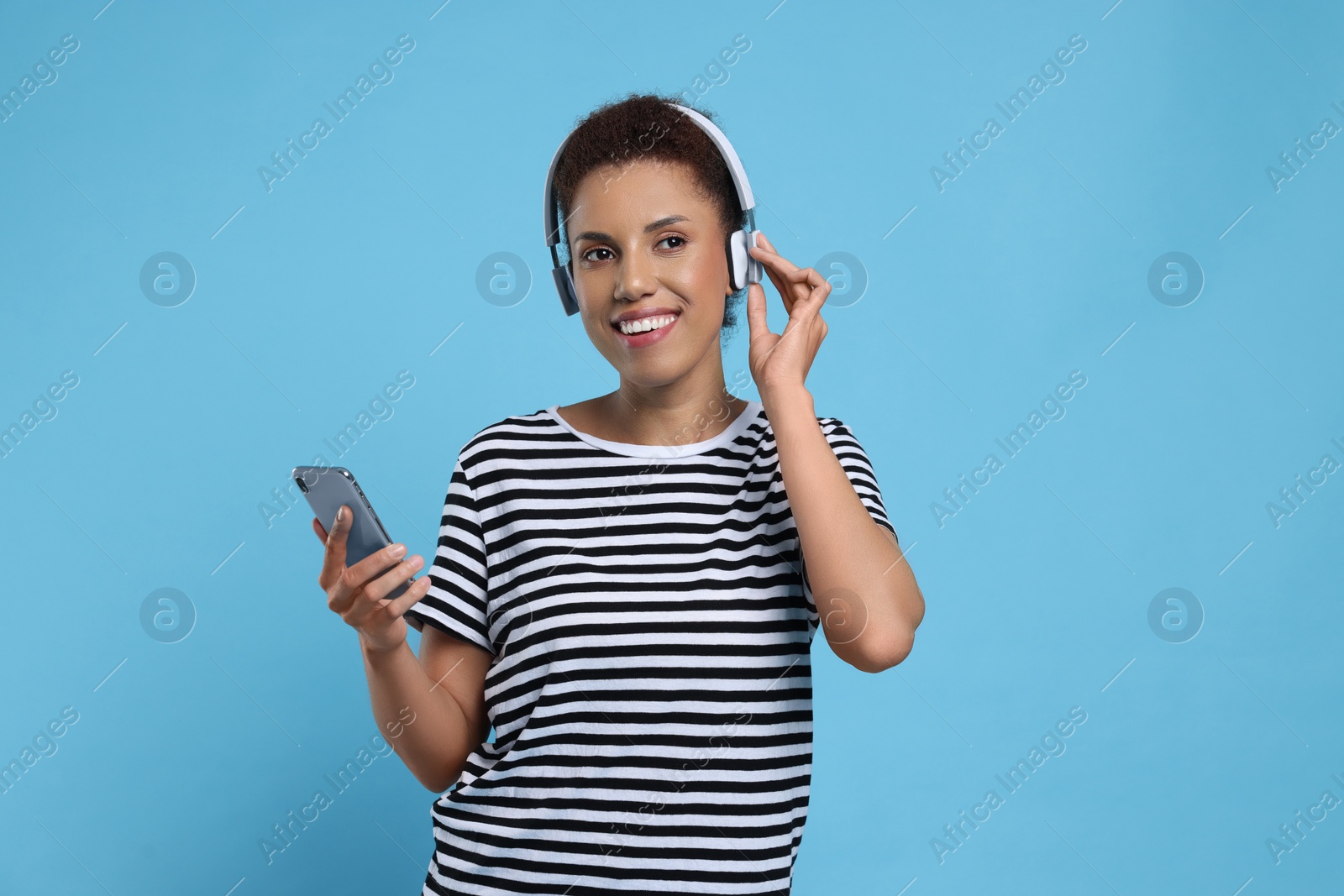 Photo of Happy young woman in headphones dancing with smartphone on light blue background