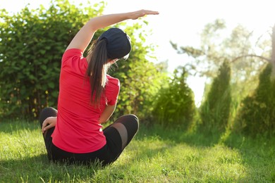 Young woman doing morning exercise on green grass in park, back view. Space for text