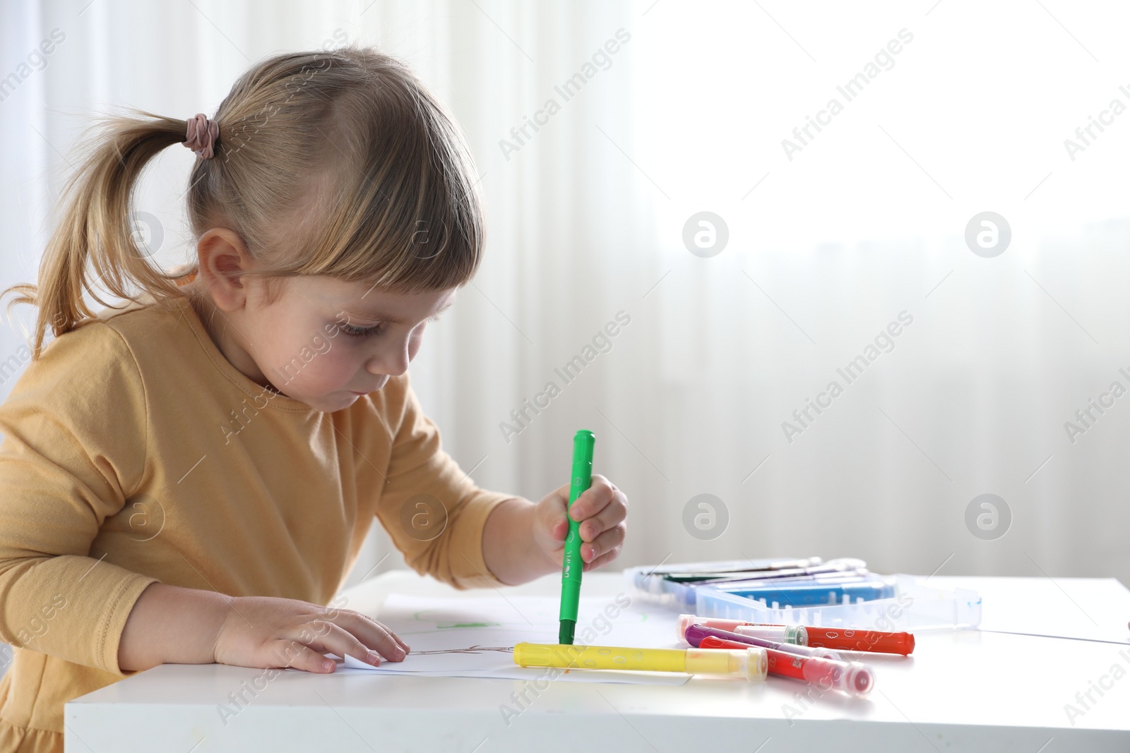 Photo of Cute little girl drawing with marker at white table indoors. Child`s art