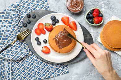 Photo of Woman eating tasty pancakes with chocolate paste and berries at grey table, top view