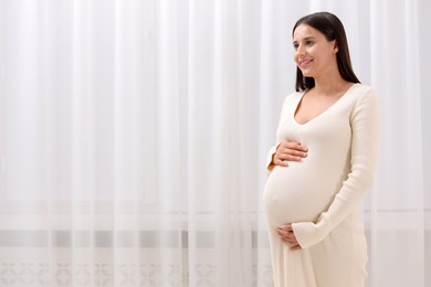 Beautiful pregnant woman in white dress indoors, space for text