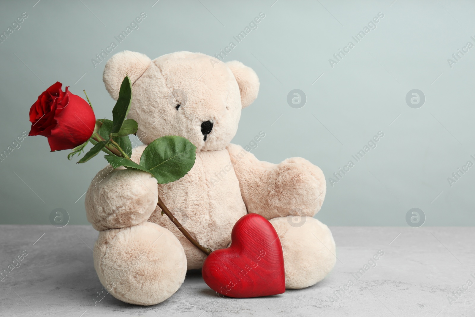 Photo of Cute teddy bear with heart and red rose on light grey stone table. Valentine's day celebration