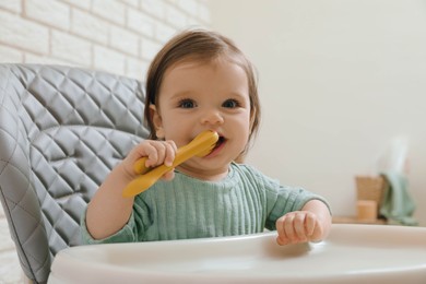 Photo of Cute little baby nibbling cutlery in high chair indoors