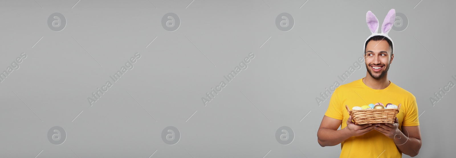 Image of Man with bunny ears holding basket full of Easter eggs on light grey background, space for text. Banner design