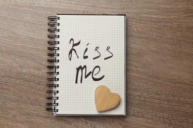 Notebook with phrase Kiss Me and decorative heart on wooden table, top view