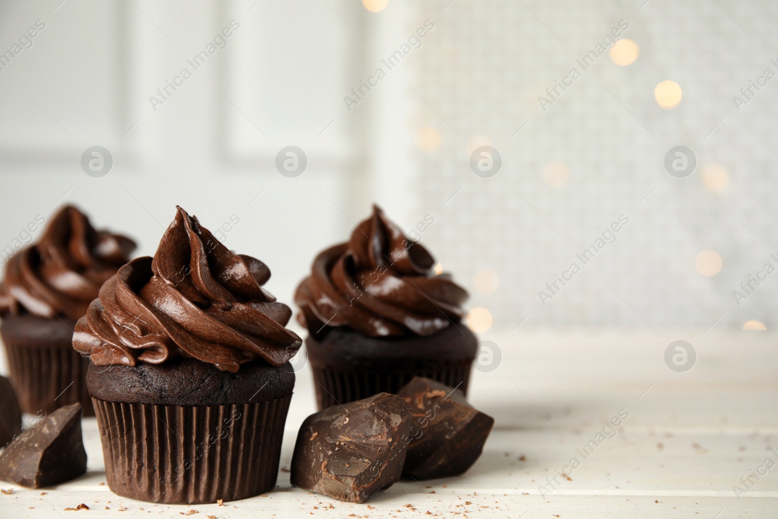 Photo of Delicious chocolate cupcakes with cream on white wooden table against blurred lights. Space for text