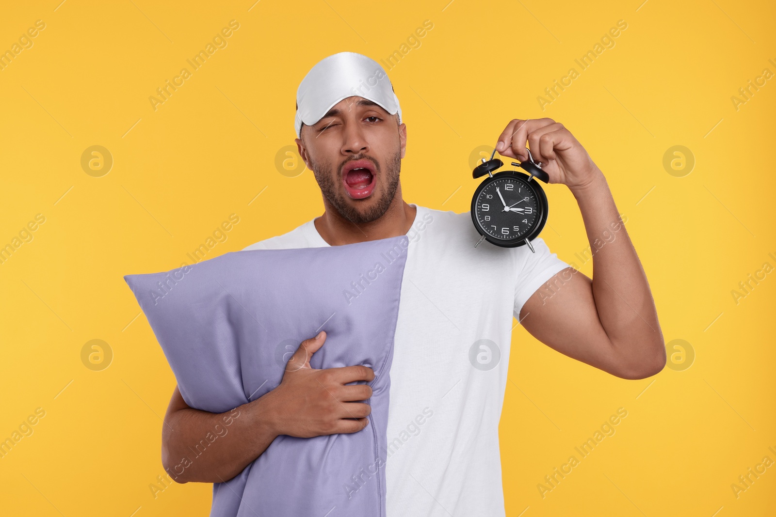 Photo of Tired man with pillow, sleep mask and alarm clock yawning on orange background. Insomnia problem