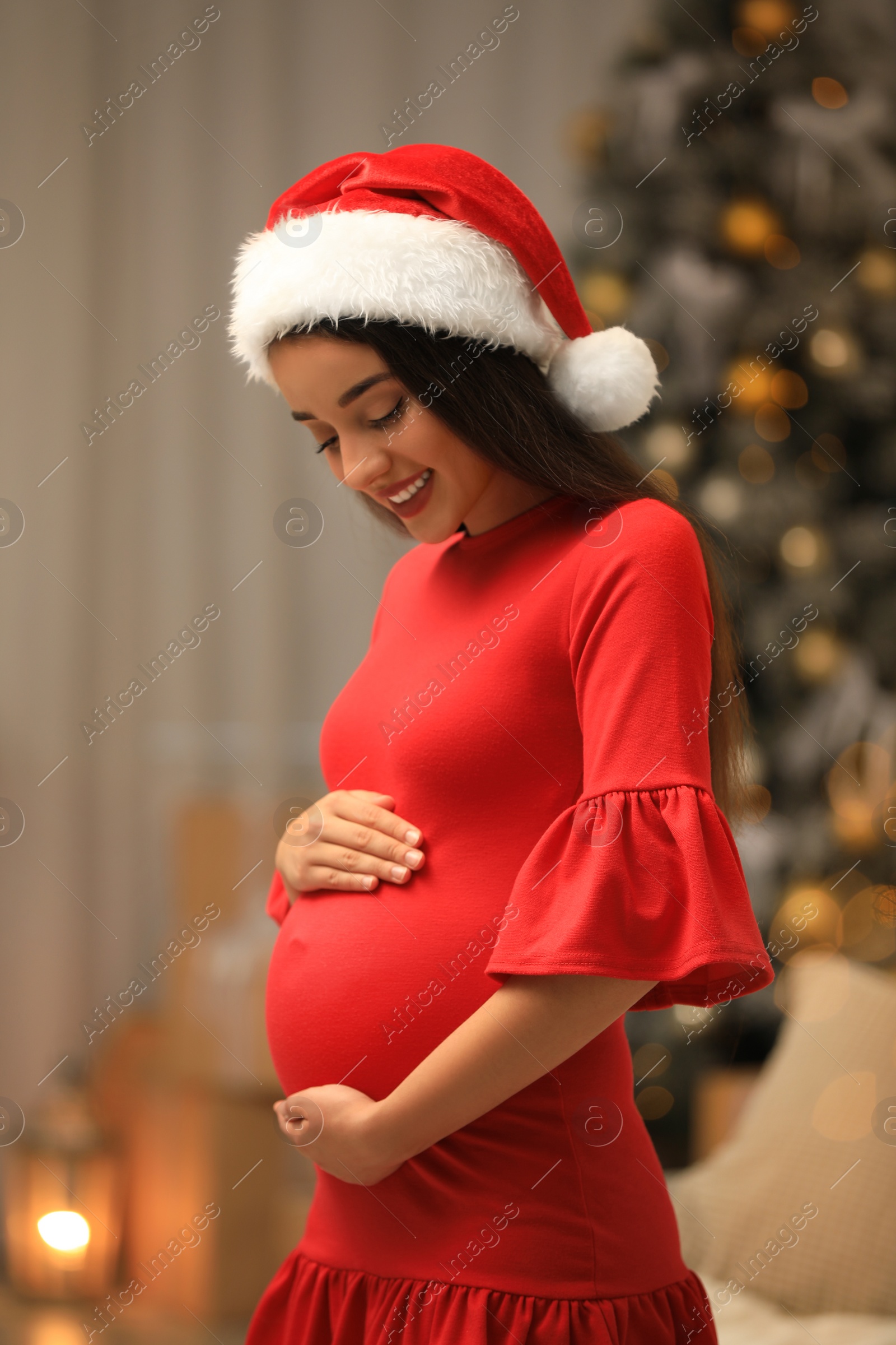 Photo of Happy pregnant woman wearing Santa hat in room decorated for Christmas. Expecting baby