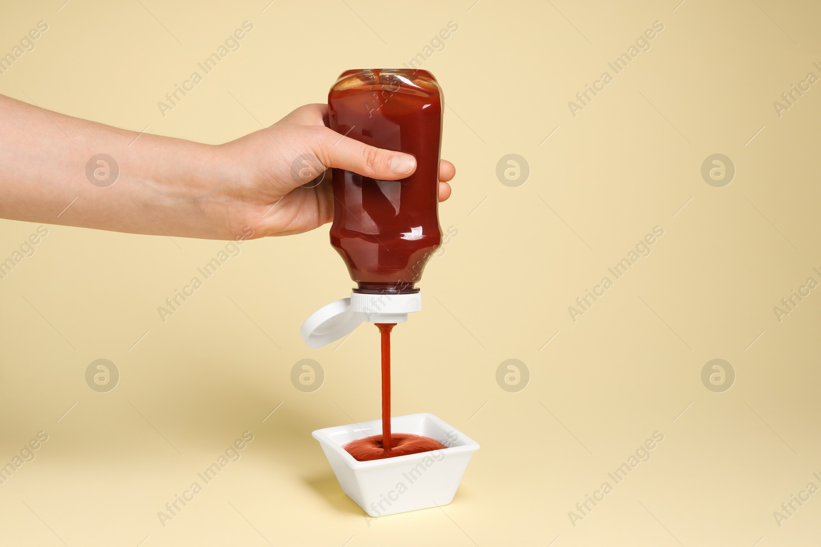 Photo of Woman pouring tasty ketchup from bottle into bowl on beige background, closeup. Space for text