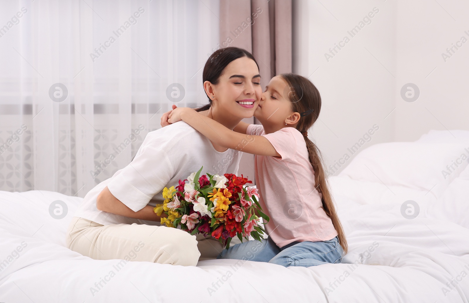 Photo of Happy woman with her daughter and bouquet of lilies on bed at home. Mother's day celebration