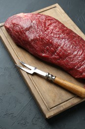 Photo of Wooden board with piece of raw meat and fork on black table