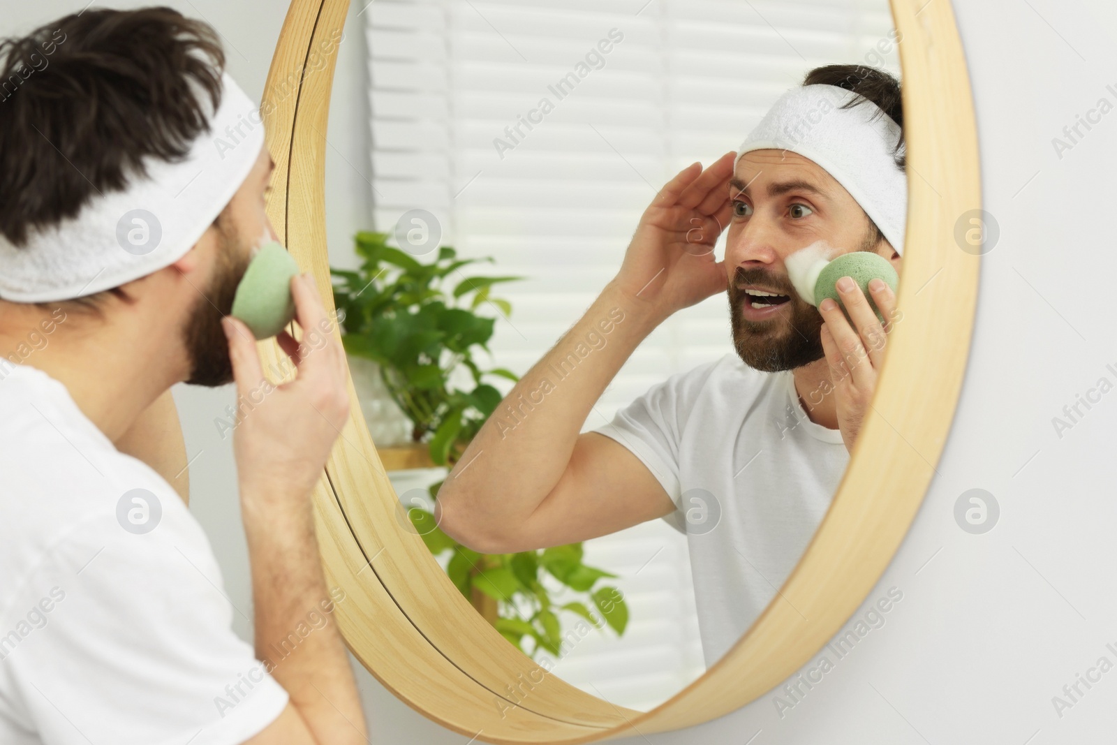 Photo of Emotional man with headband washing his face using sponge near mirror in bathroom