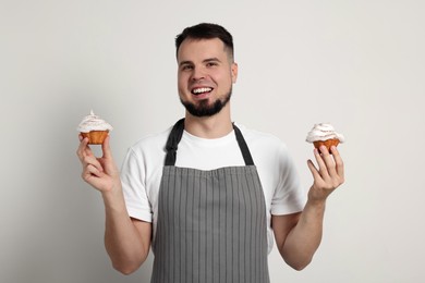 Happy professional confectioner in apron holding delicious cupcakes on light grey background