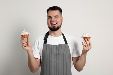 Photo of Happy professional confectioner in apron holding delicious cupcakes on light grey background