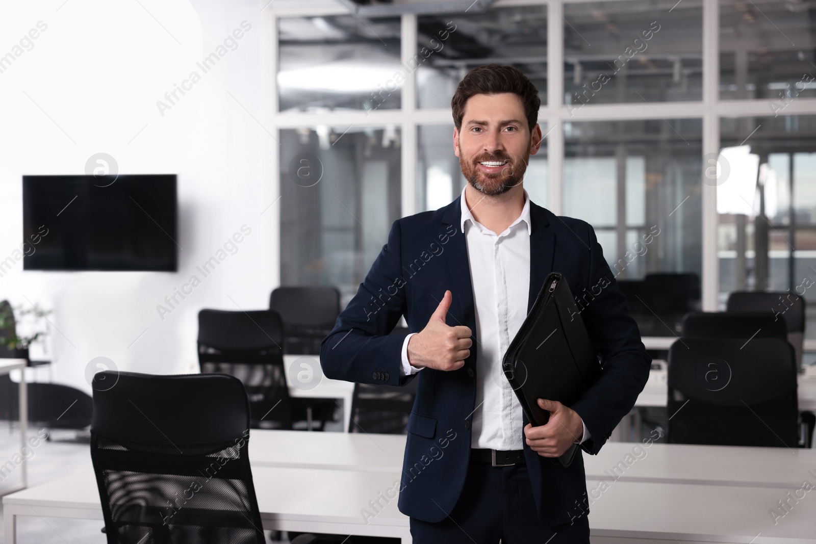 Photo of Happy real estate agent with leather portfolio showing thumb up indoors