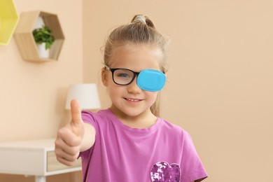 Girl with eye patch on glasses showing thumb up in room. Strabismus treatment