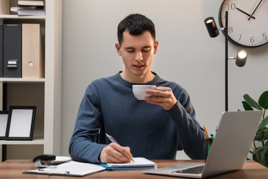 Photo of Man with cup of drink taking notes at wooden table in office