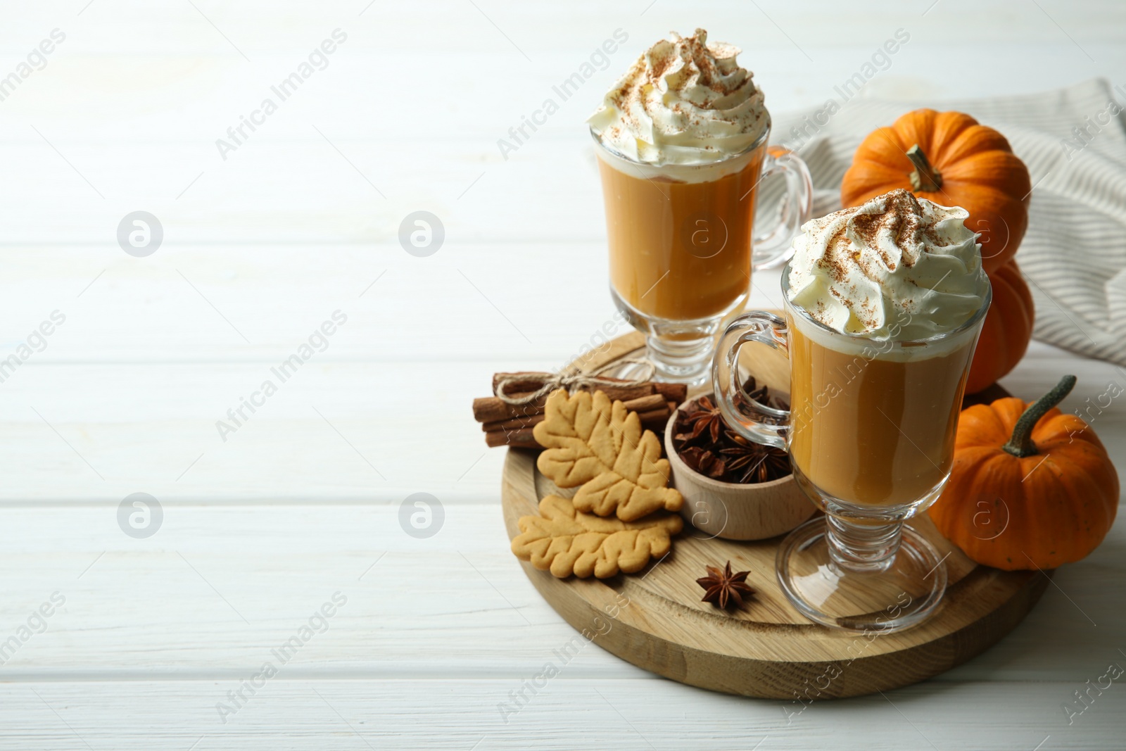 Photo of Tasty pumpkin latte with whipped cream in glasses, spices and cookies on white wooden table. Space for text