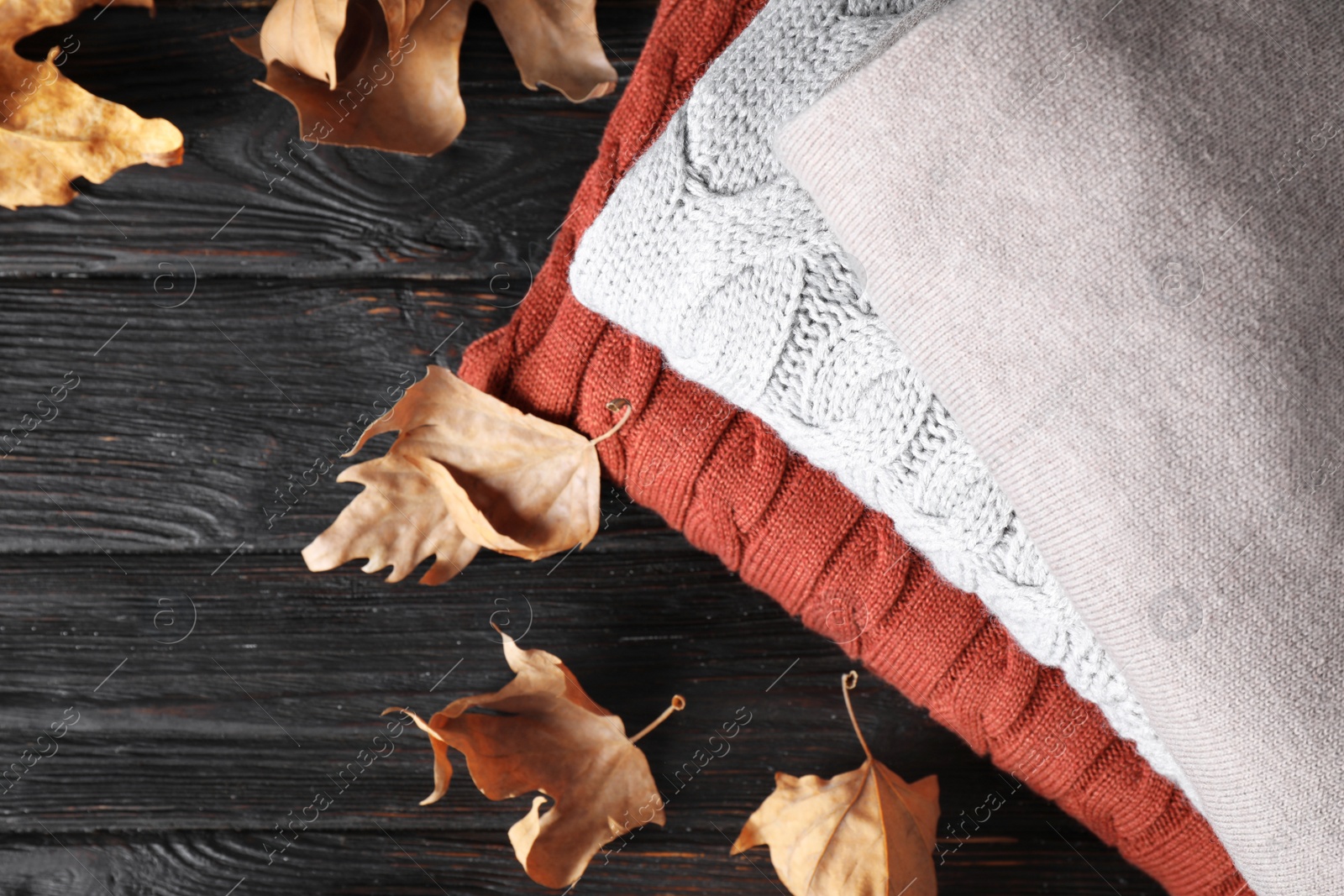 Image of Stack of folded warm sweaters and dry leaves on black wooden table, top view