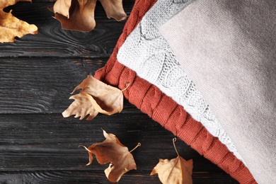 Stack of folded warm sweaters and dry leaves on black wooden table, top view