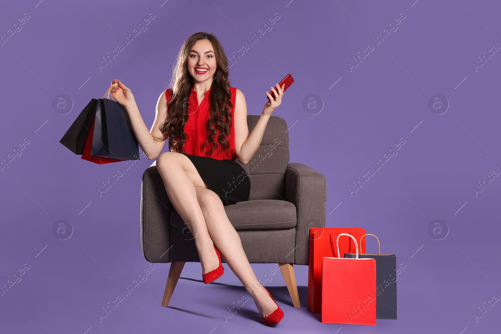 Photo of Happy woman holding paper shopping bags and smartphone on armchair against purple background