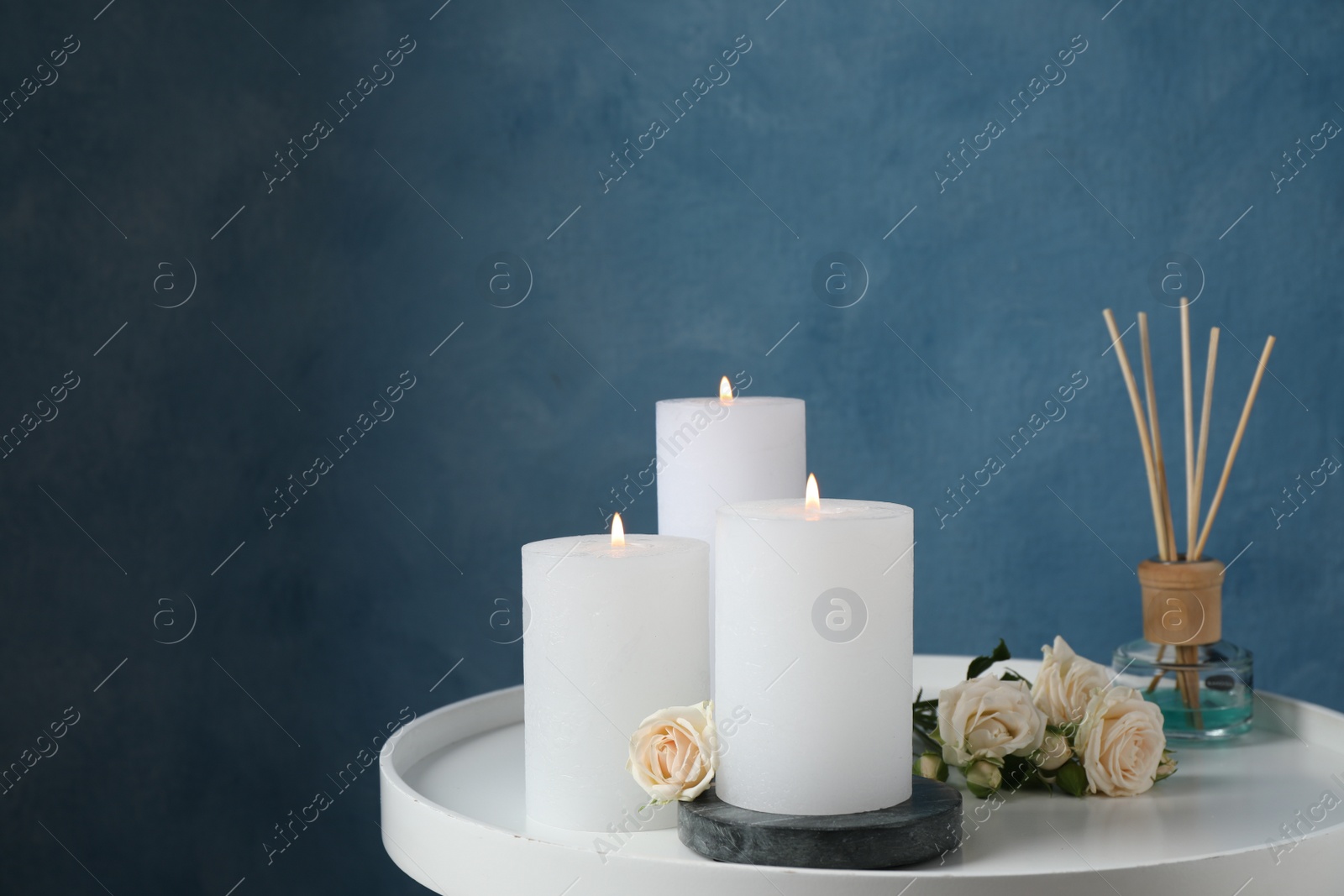 Photo of Burning candles, air reed diffuser and beautiful roses on white table near light blue wall, space for text