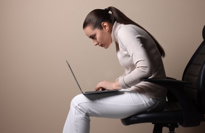 Young woman with poor posture using laptop while sitting on chair against beige background