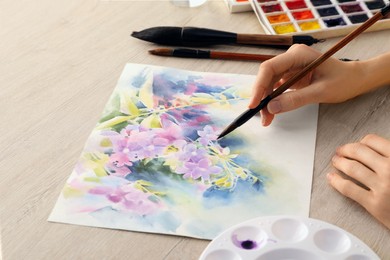 Photo of Woman painting flowers with watercolor at white wooden table, closeup. Creative artwork