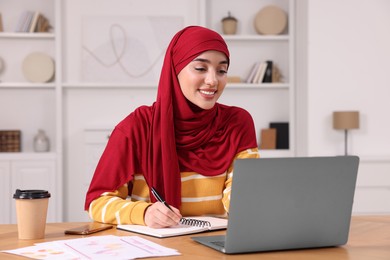 Photo of Muslim woman writing notes near laptop at wooden table at home