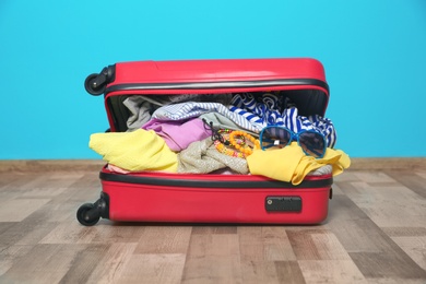 Photo of Open suitcase with different clothes and accessories packed for summer journey on floor near color wall