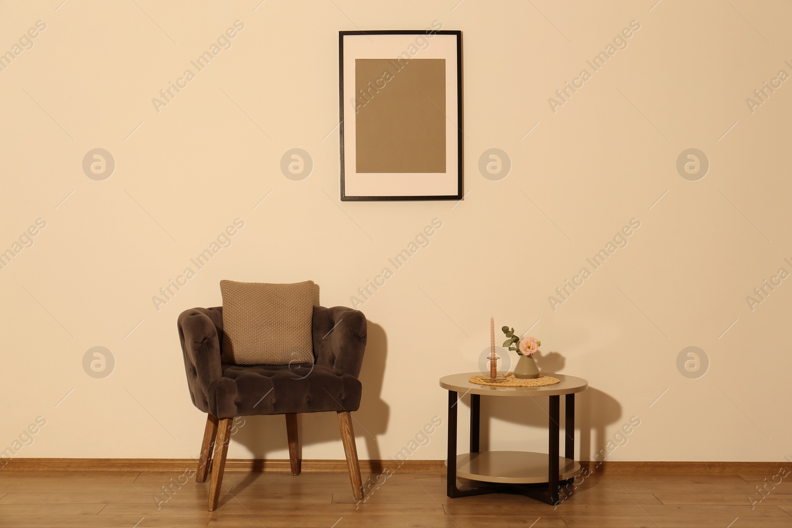 Photo of Comfortable armchair, cushion, table and decor elements in room with beige wall. Interior design