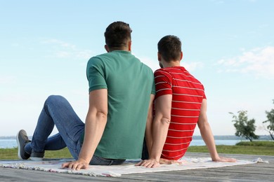 Photo of Gay couple sitting near river on sunny day, back view