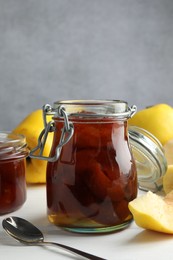 Tasty homemade quince jam in jar, spoon and fruits on white wooden table, closeup