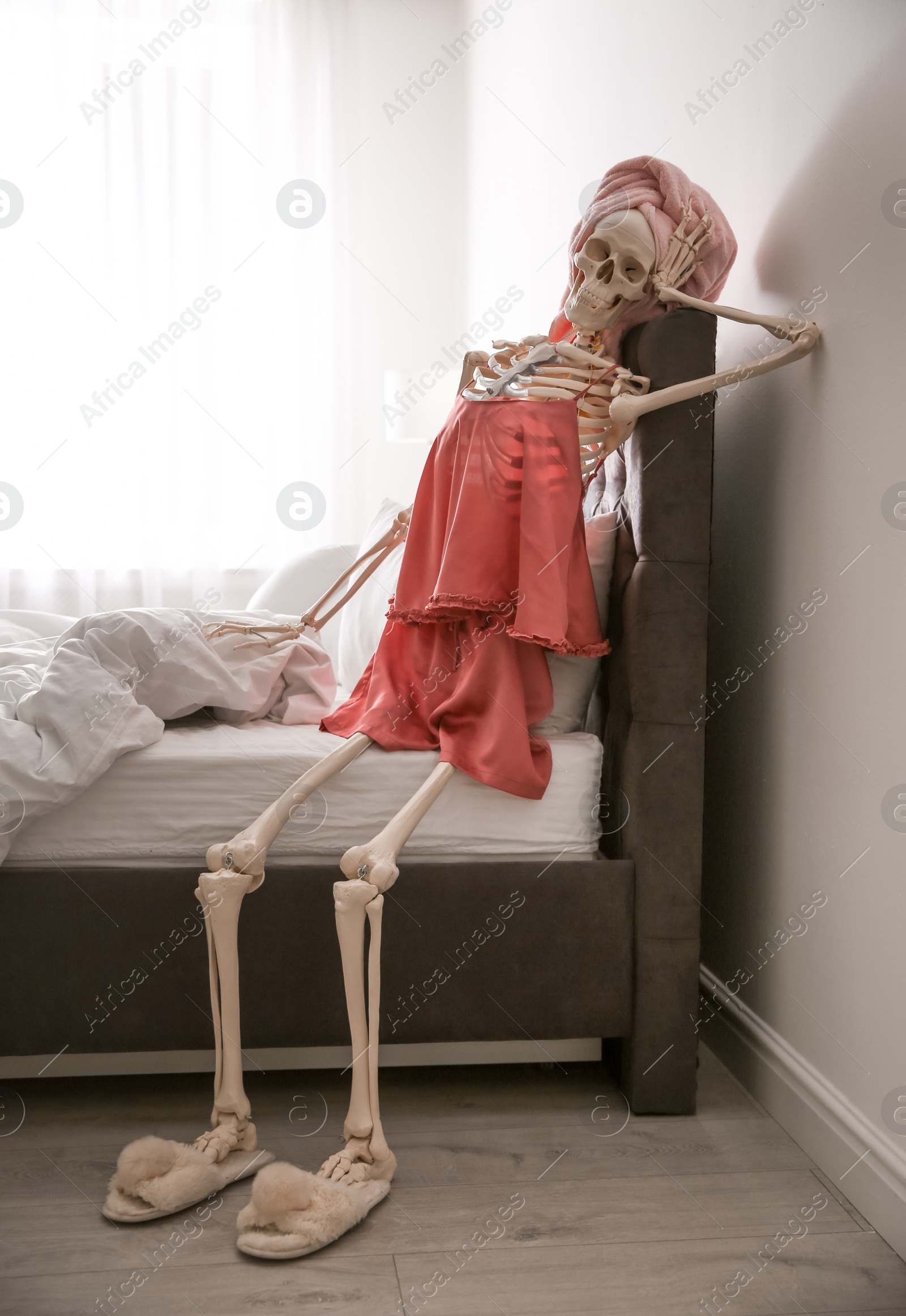 Photo of Human skeleton in silk pajamas and towel sitting on bed indoors