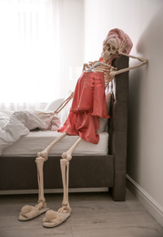 Photo of Human skeleton in silk pajamas and towel sitting on bed indoors