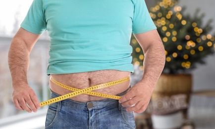 Overweight man measuring his waist in room decorated for Christmas after holidays, closeup