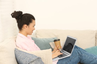 African American woman with laptop and coffee on sofa in room