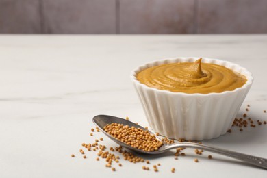 Photo of Tasty mustard sauce in bowl and spoon with dry seeds on white marble table, space for text
