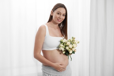 Beautiful pregnant woman with bouquet of roses near window indoors