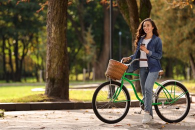 Young woman with bicycle holding takeaway coffee in park, space for text