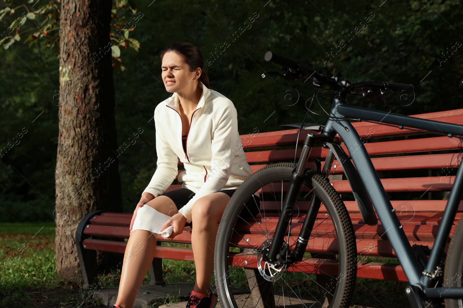 Photo of Young woman applying bandage onto her knee on wooden bench outdoors