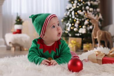 Photo of Baby wearing cute elf costume on floor in room decorated for Christmas