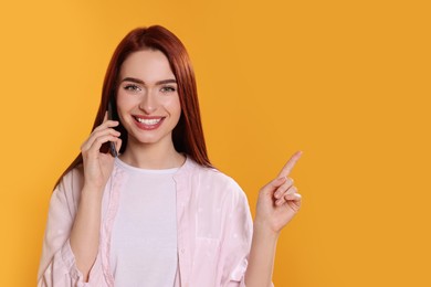 Photo of Happy woman with red dyed hair talking on phone and pointing somewhere against orange background, space for text