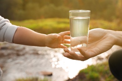 Photo of Mother giving her daughter glass of fresh water near stream on sunny day, closeup