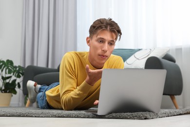 Photo of Happy young man having video chat via laptop and blowing kiss on carpet indoors. Long-distance relationship