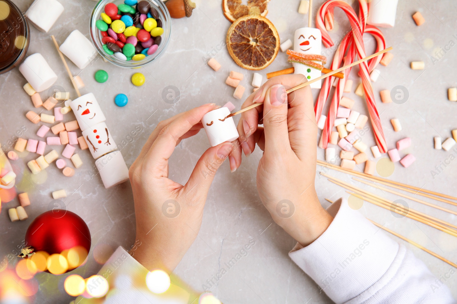 Image of Woman making funny snowman with marshmallows at light marble table, top view. Bokeh effect 