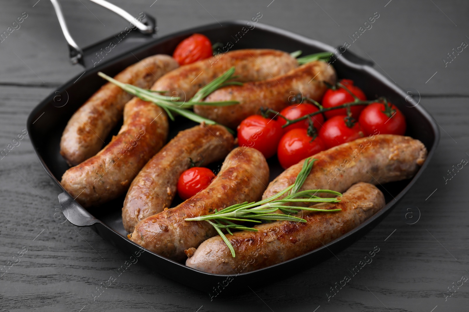 Photo of Grill pan with tasty homemade sausages, rosemary and tomatoes on grey wooden table, closeup