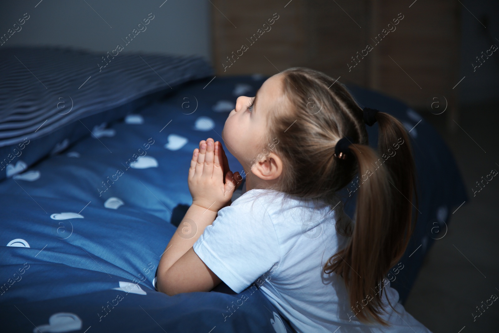 Photo of Little girl saying bedtime prayer near bed in room at night