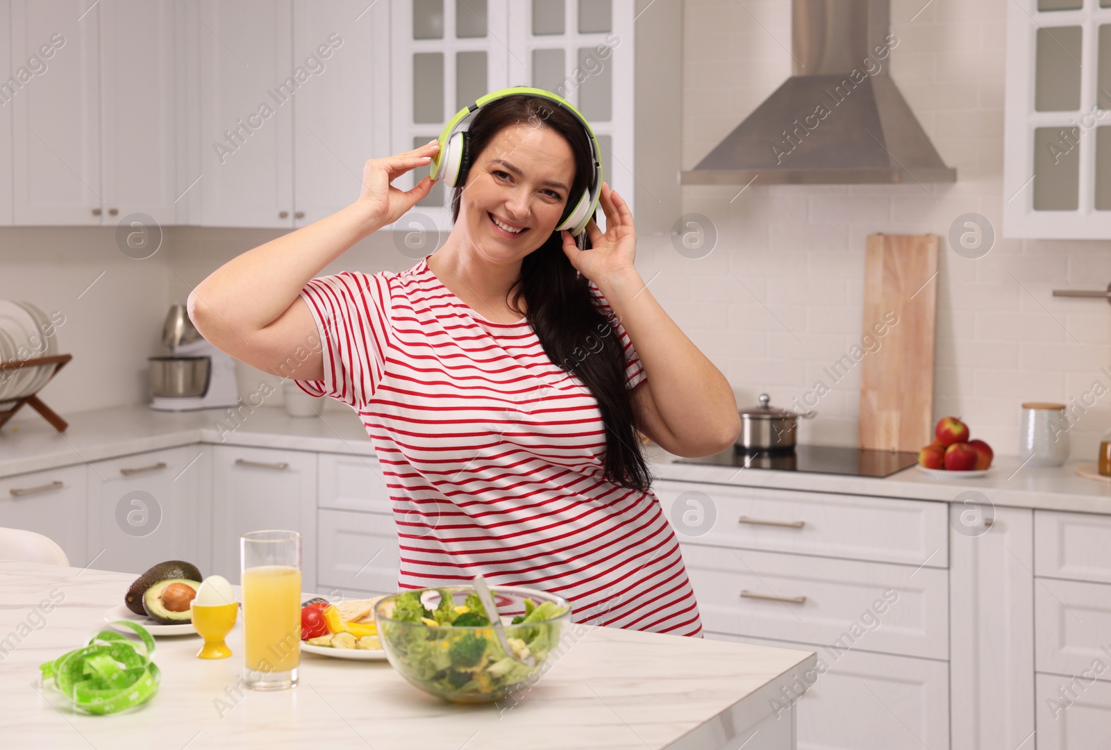 Photo of Happy overweight woman with headphones dancing near table in kitchen. Healthy diet