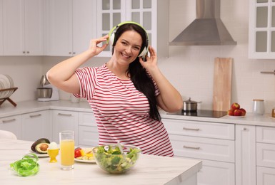 Happy overweight woman with headphones dancing near table in kitchen. Healthy diet